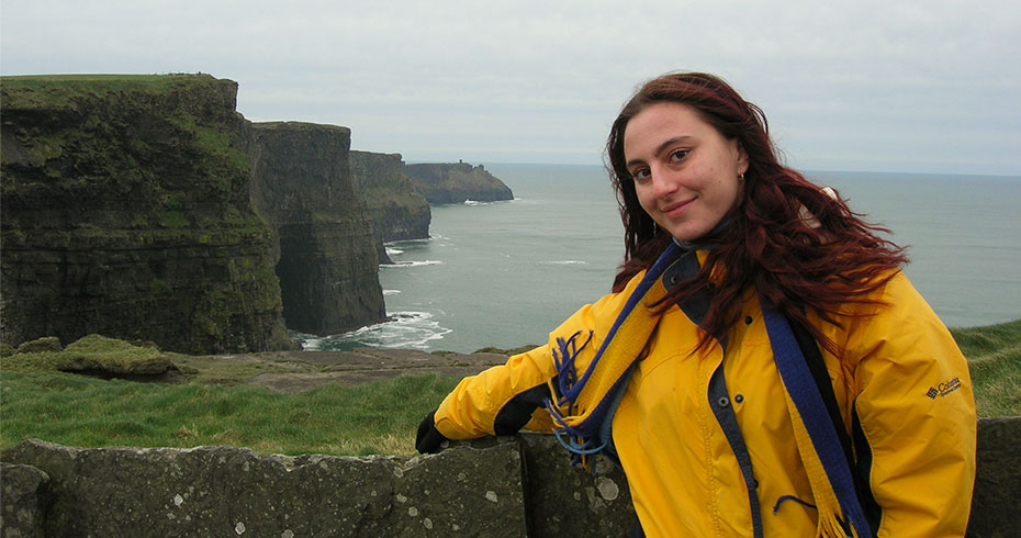 A young woman wearing a bright yellow jacket poses in front of the Cliffs of Moher on a cloudy day.
