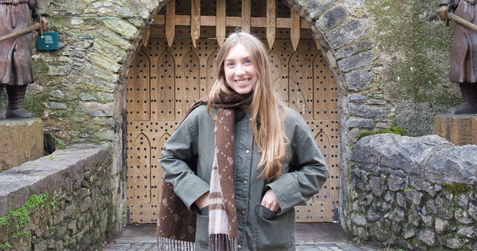 A young woman with blonde hair stands in front of a medieval castle. She is wearing a khaki jacket and a brown scarf.
