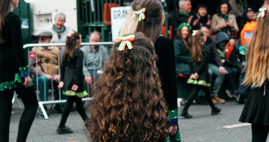 Young dancers wearing black costumes with green details and tricolour hair ribbons perform during a St Patrick’s Day parade in Ireland, as spectators watch from the roadside at a lively Irish festival celebration.