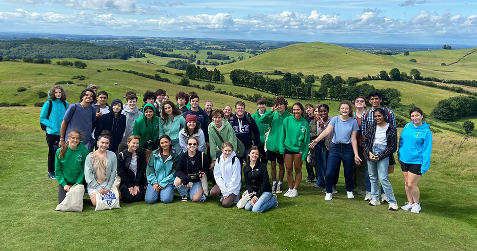 A group of international students on a high school summer programme at UCD. They are posing and smiling on a green hillside under a blue sky.